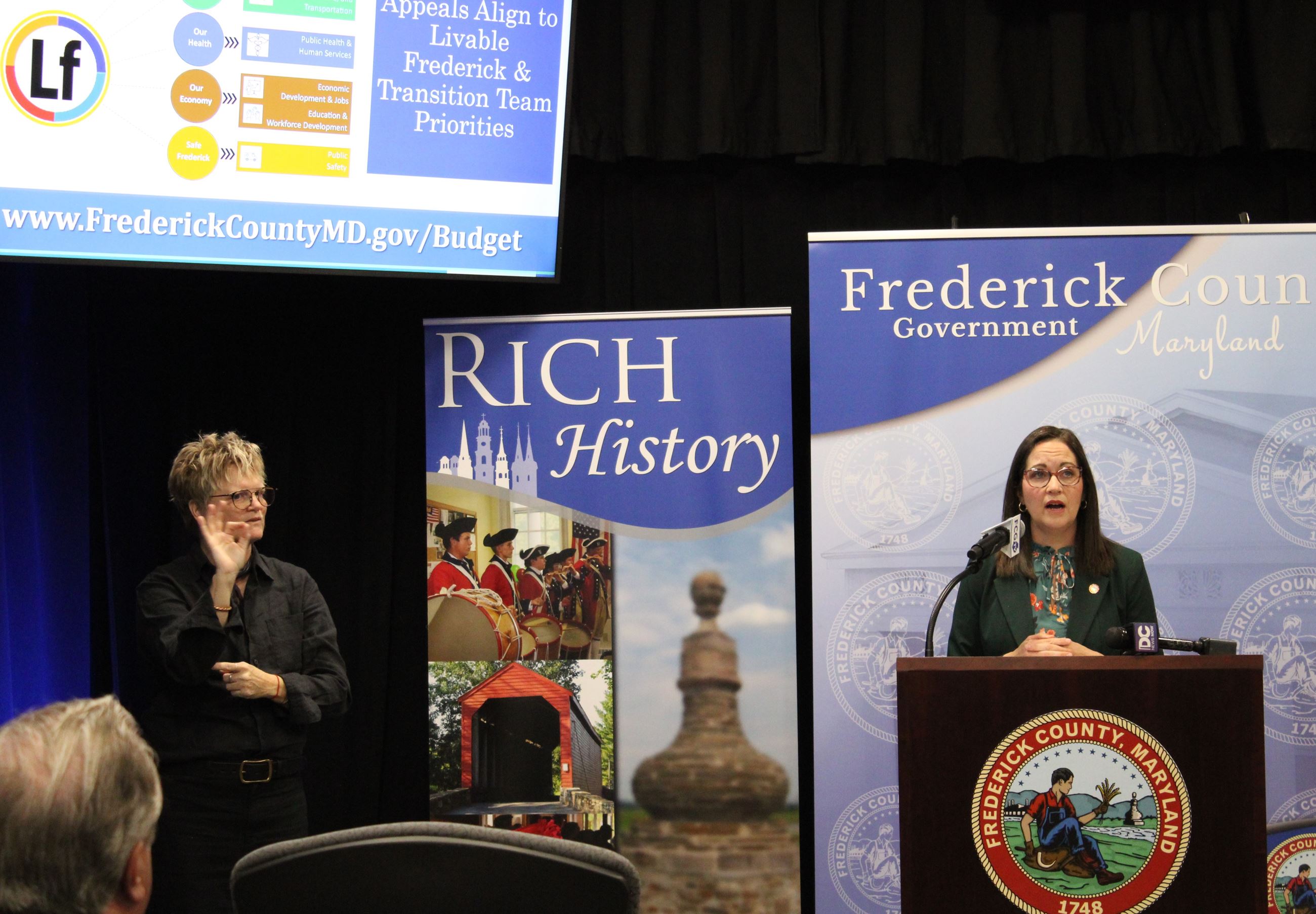 Two people stand inside, one of them behind a lectern with a Frederick County seal on it.